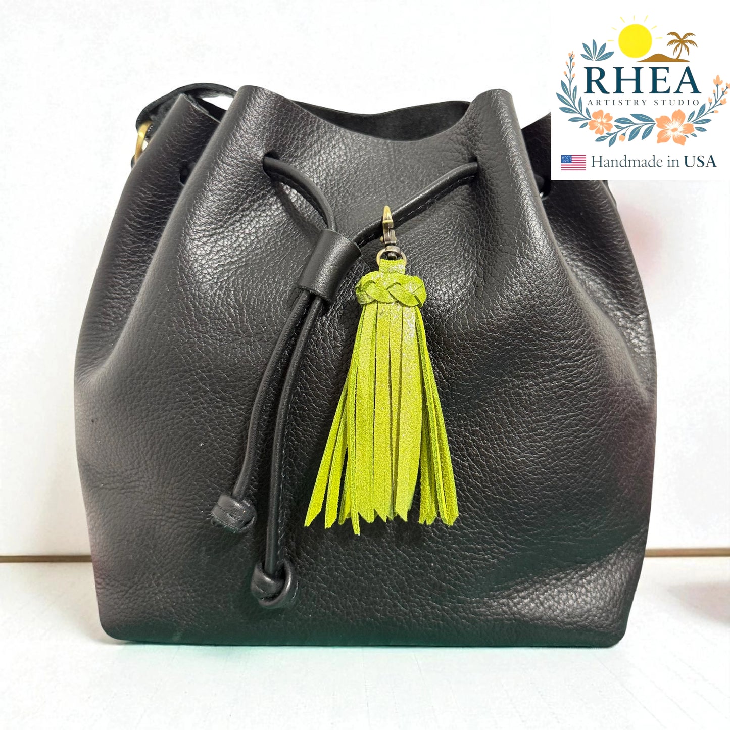 Black leather bucket bag with a green tassel on a white background, featuring the RHEA brand logo.