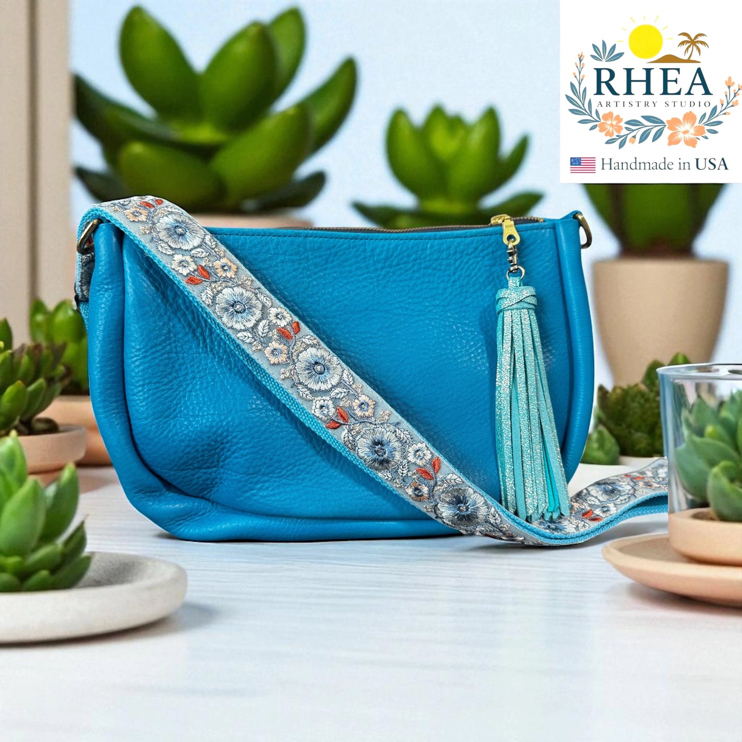 Blue handbag with decorative fabric artisan strap on a light surface with plants in the background, featuring the RHEEA brand.