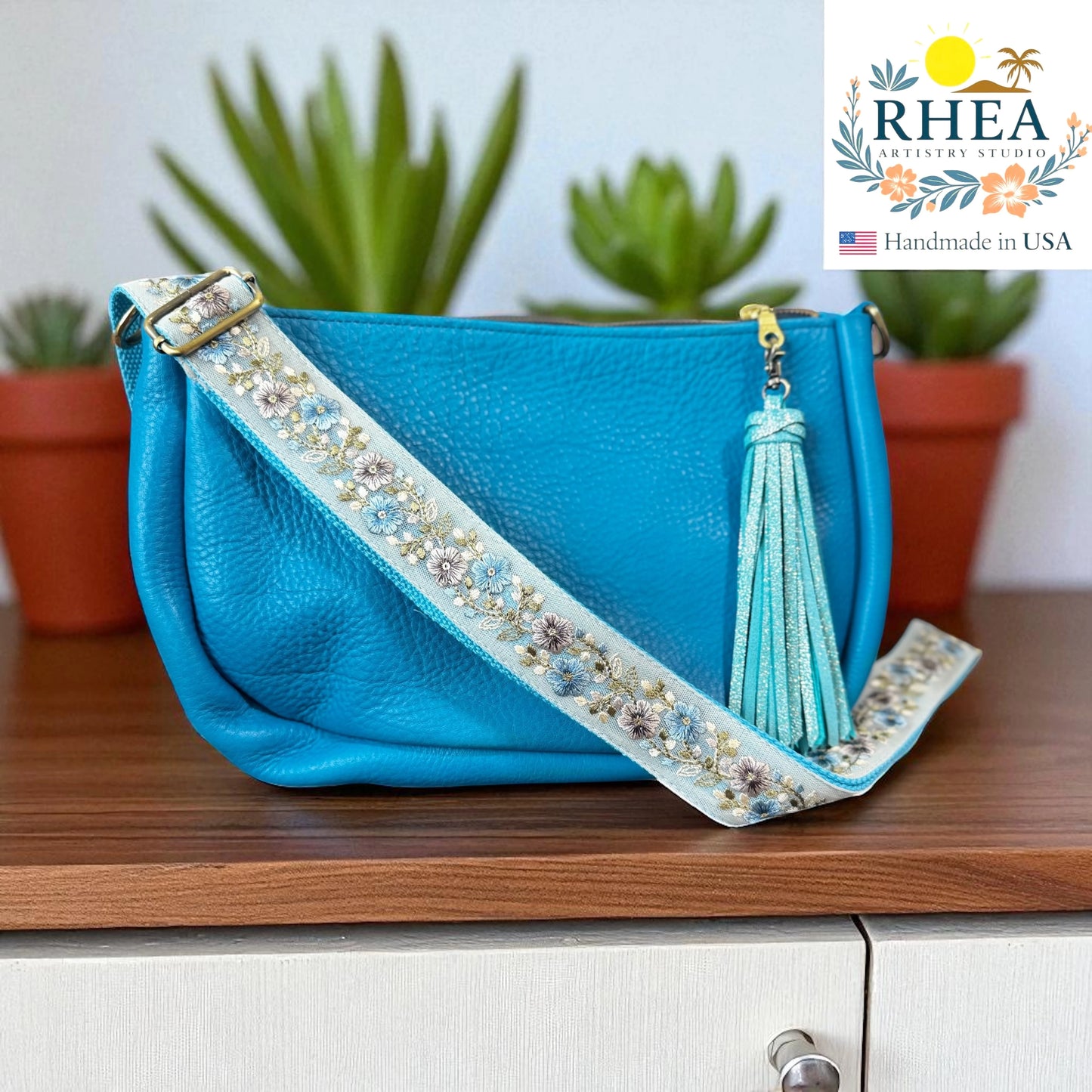 Blue handbag with decorative embroidered crossbody bag strap and tassel on a wooden surface, RHEA brand visible.
