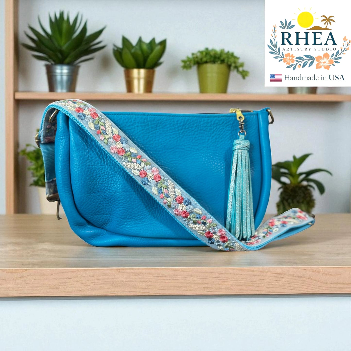 Blue handbag with floral artisan bag strap on a wooden surface, RHEA brand visible