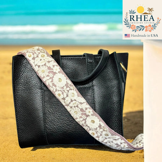 Black handbag with a floral purse strap on a beach, featuring the RHEA brand.