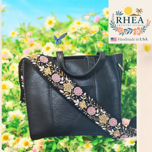 Black handbag with floral embroidery against a colorful floral background, featuring the Rhea Artisan Studio logo.