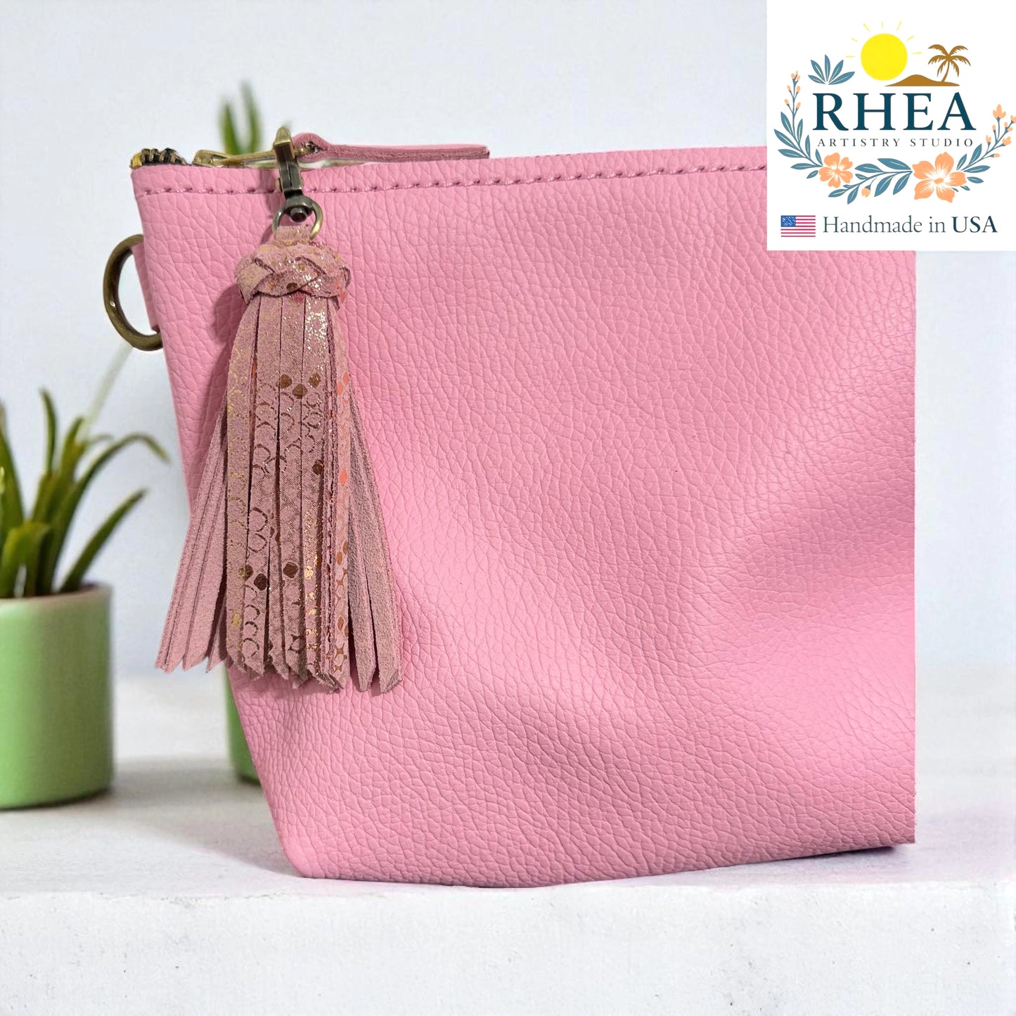 Pink pouch with a leather bag tassel on a white surface, RHEA brand visible