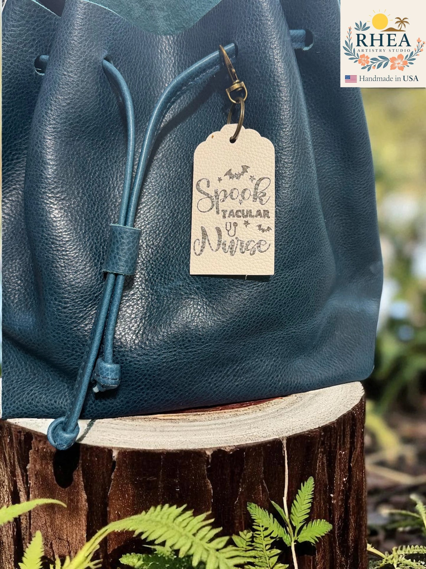 Blue leather bag with a leather engraved tag on a wooden stump with plants in the background, featuring the RHEEA brand.