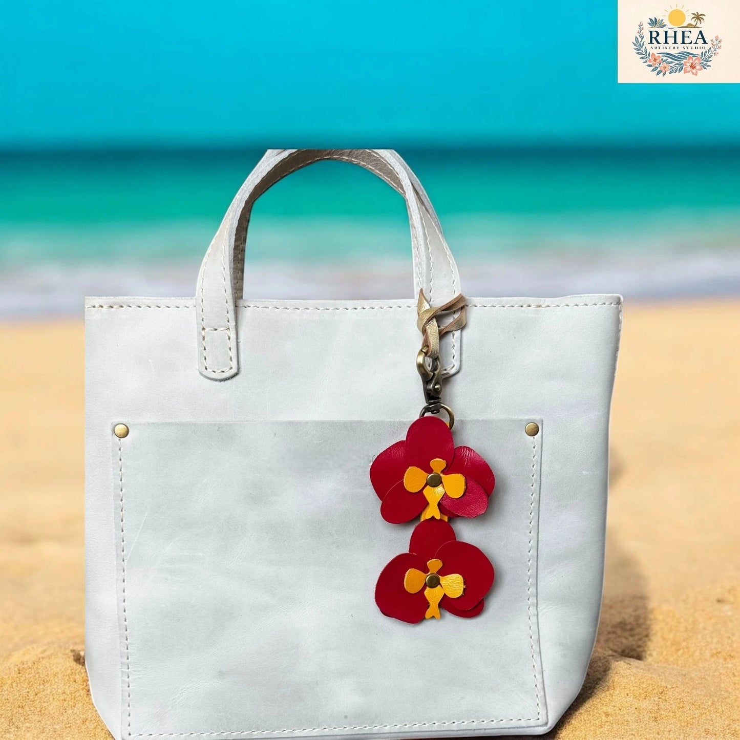 White handbag with a red floral keychain on a sandy beach with blue water in the background, featuring the RHEA brand.