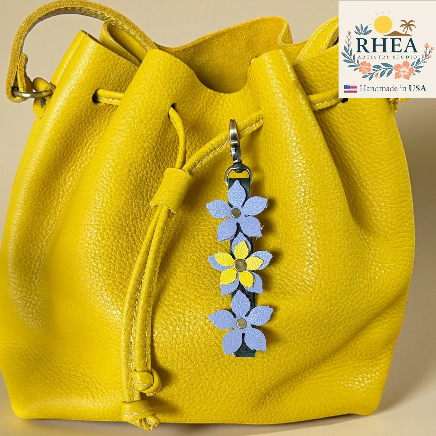 Yellow leather bag with a floral keychain charm on a beige background, featuring the RHEA Artisan Studio logo.