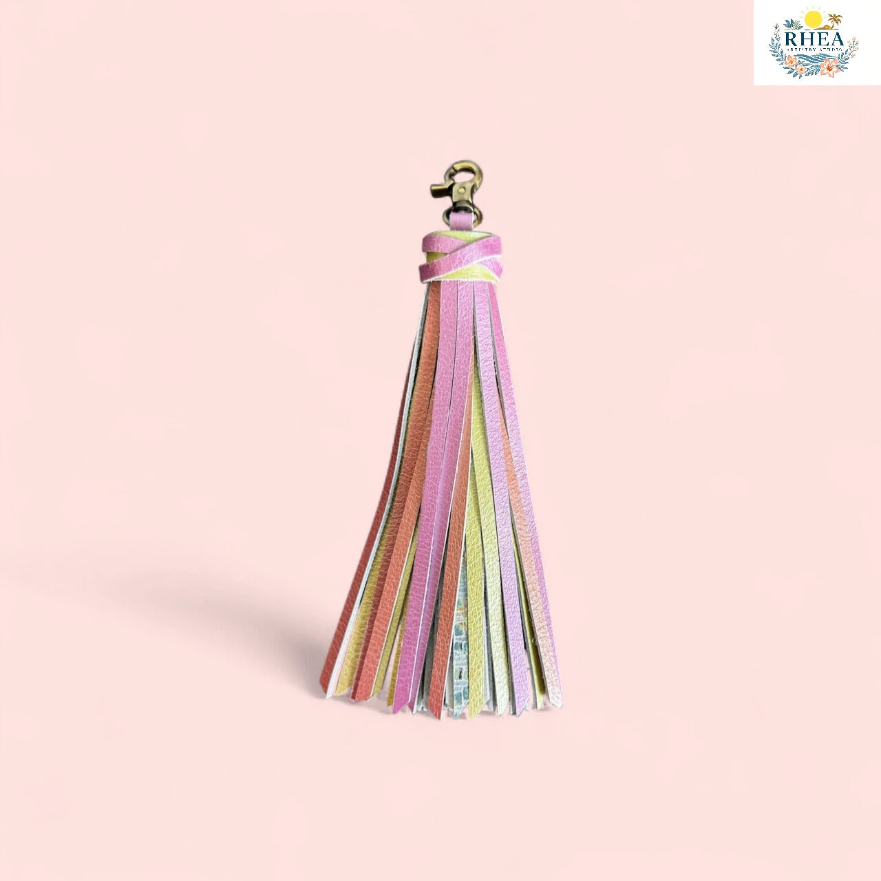 Colorful tassel keychain on a pink background with RHEA logo.