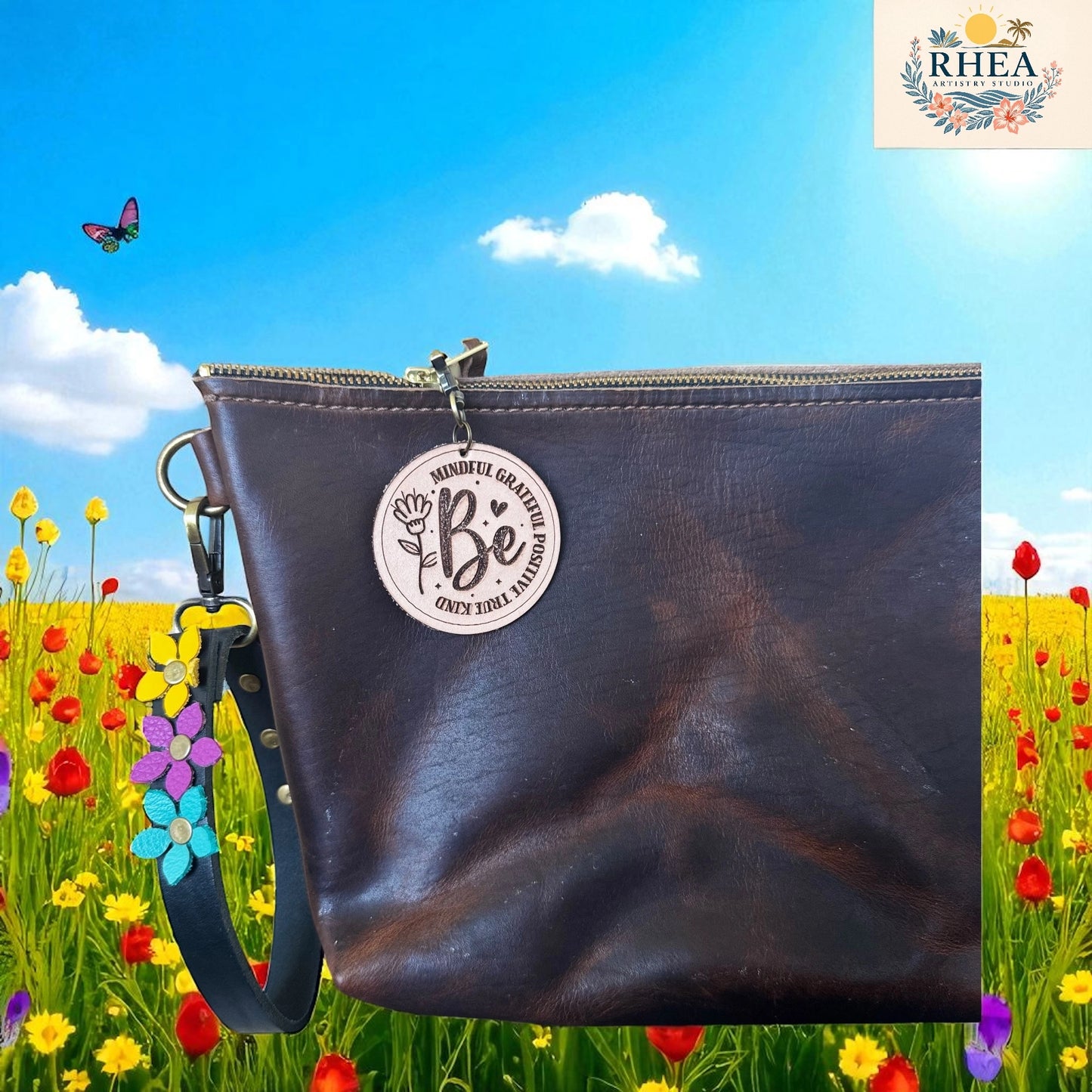 Brown leather clutch bag with 'Be' charm in a field of flowers and butterflies, featuring the RHEEA brand.