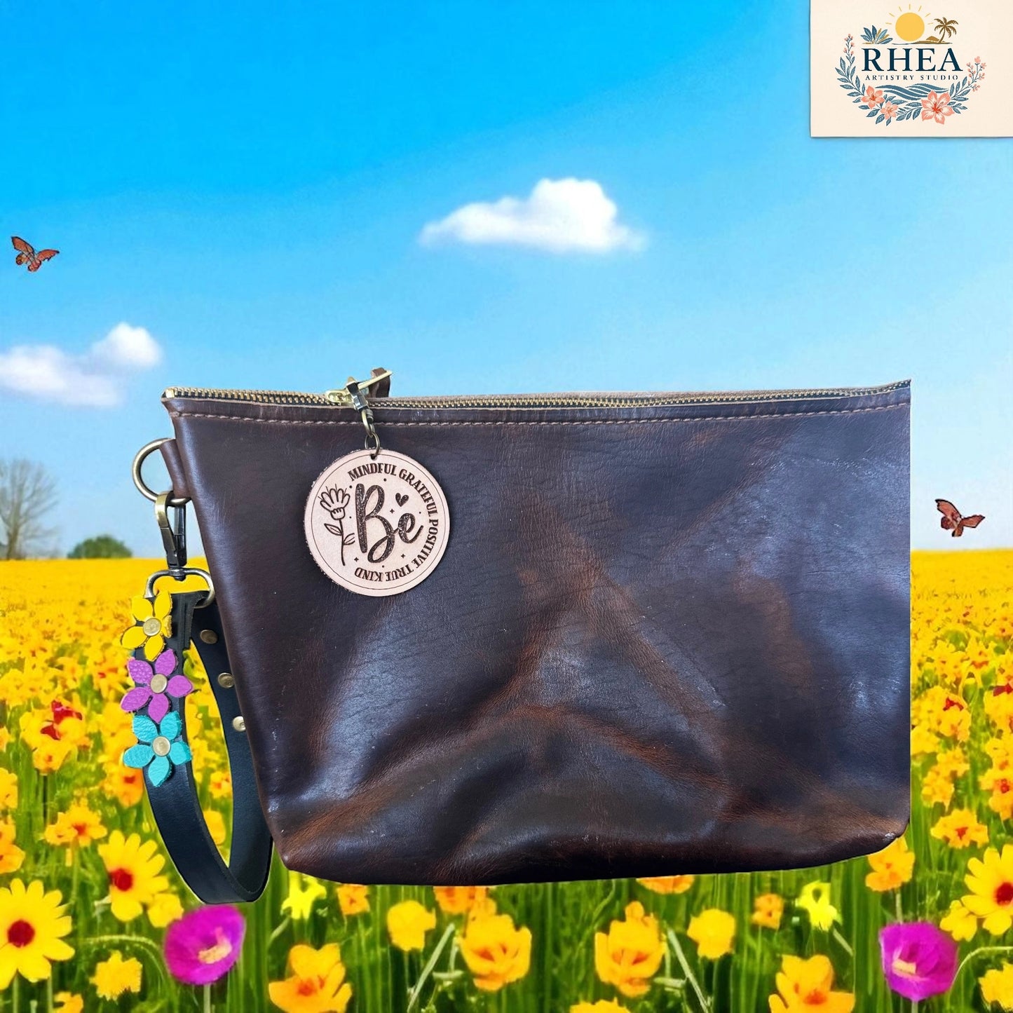 Brown leather pouch with 'Be' charm on a background of a blue sky and yellow flowers.