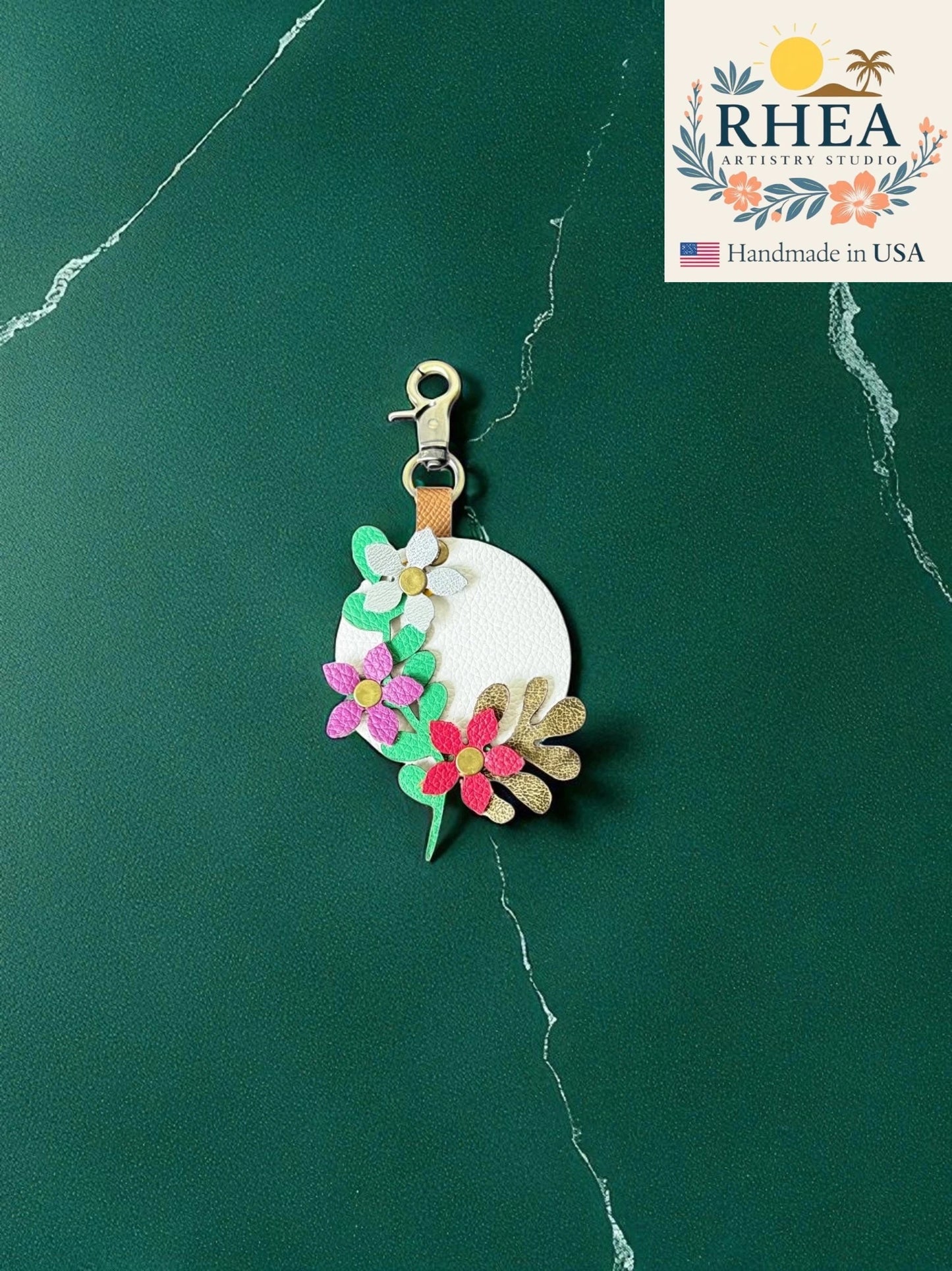 Decorative leather bag charm with floral design on a green marble background, featuring the RHEA brand.