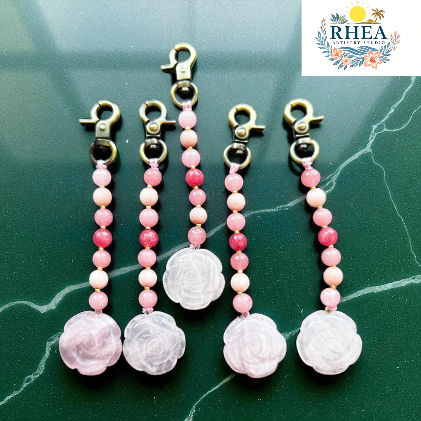 Pink beaded keychains with floral charms on a green marble surface, branded 'RHEA'.