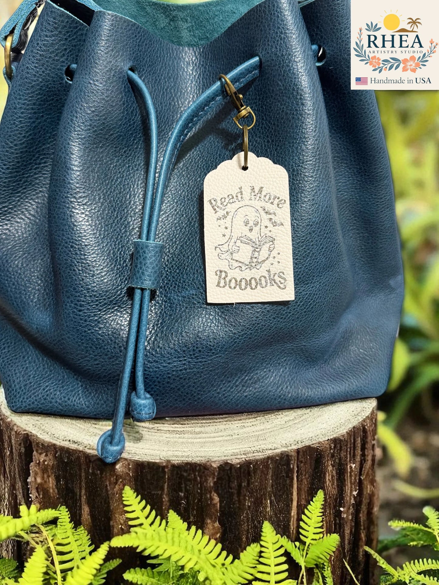 Blue leather bag with a tag on a wooden stump surrounded by greenery, featuring the RHEEA brand.