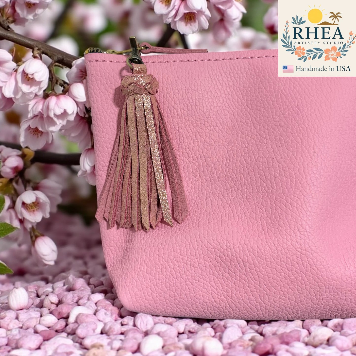 Pink handbag with a tassel detail in front of cherry blossoms, featuring the RHEA Artistry Studio logo.