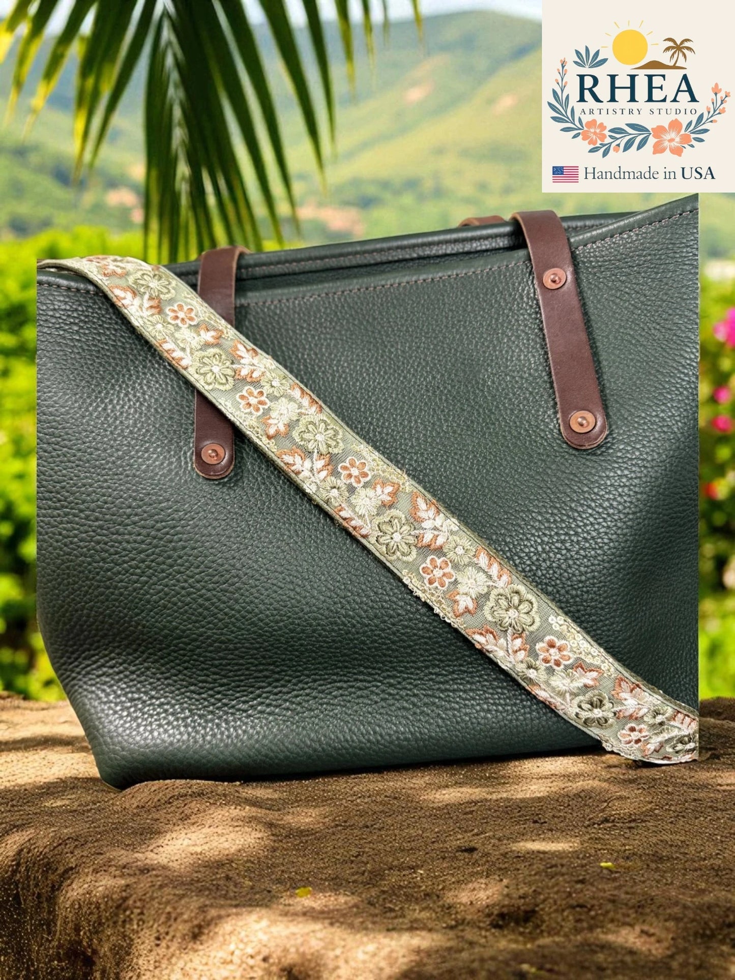 Green handbag with a floral strap on a rock with a scenic background, featuring the RHEA brand.