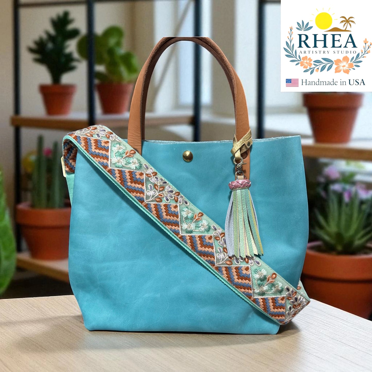Turquoise handbag with decorative strap and tassel on a wooden surface, with 'RHEA' branding.