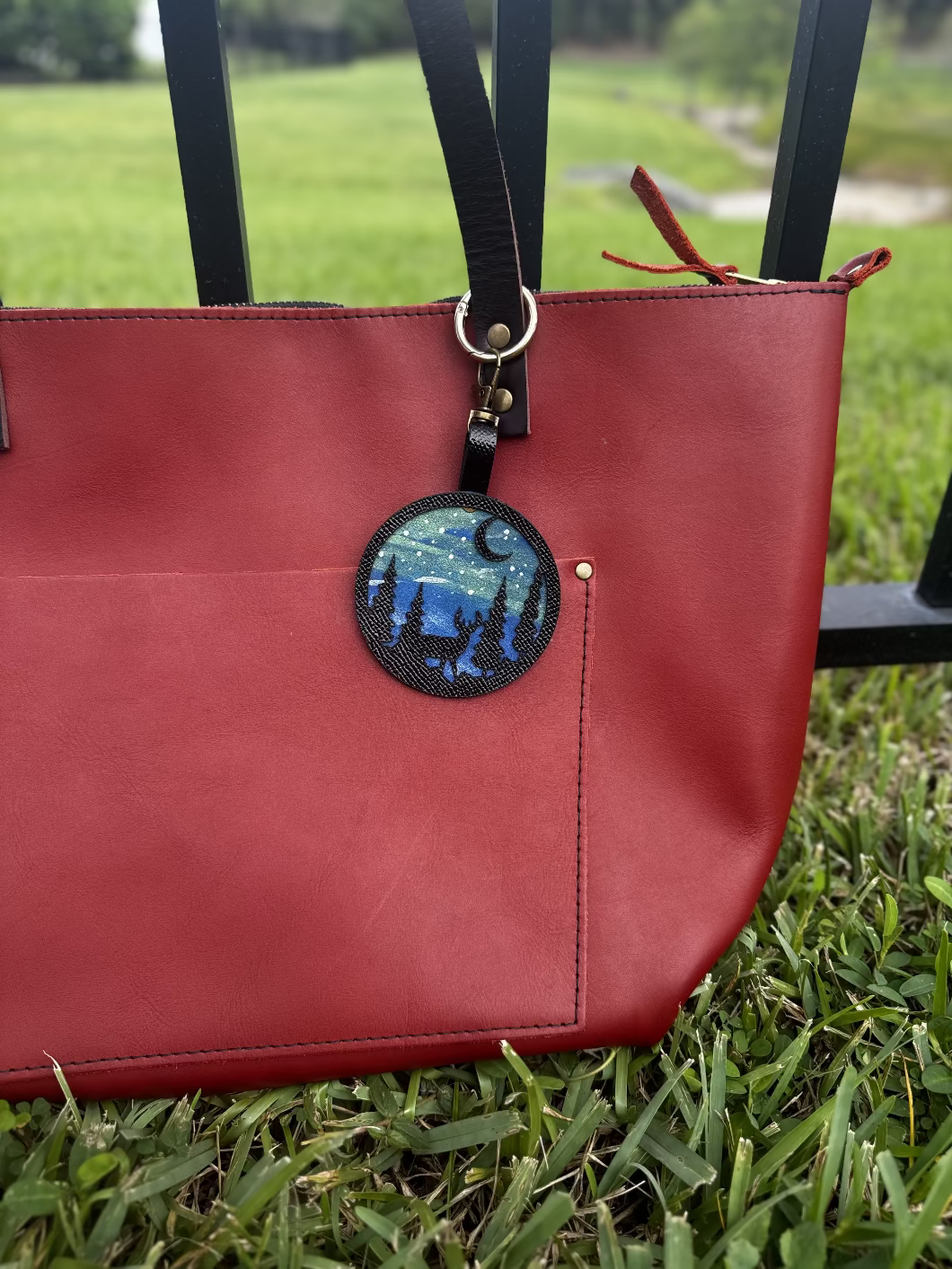 A handpainted leather charm with a starry night landscape, a deer, and a forest design, attached to a red leather bag.