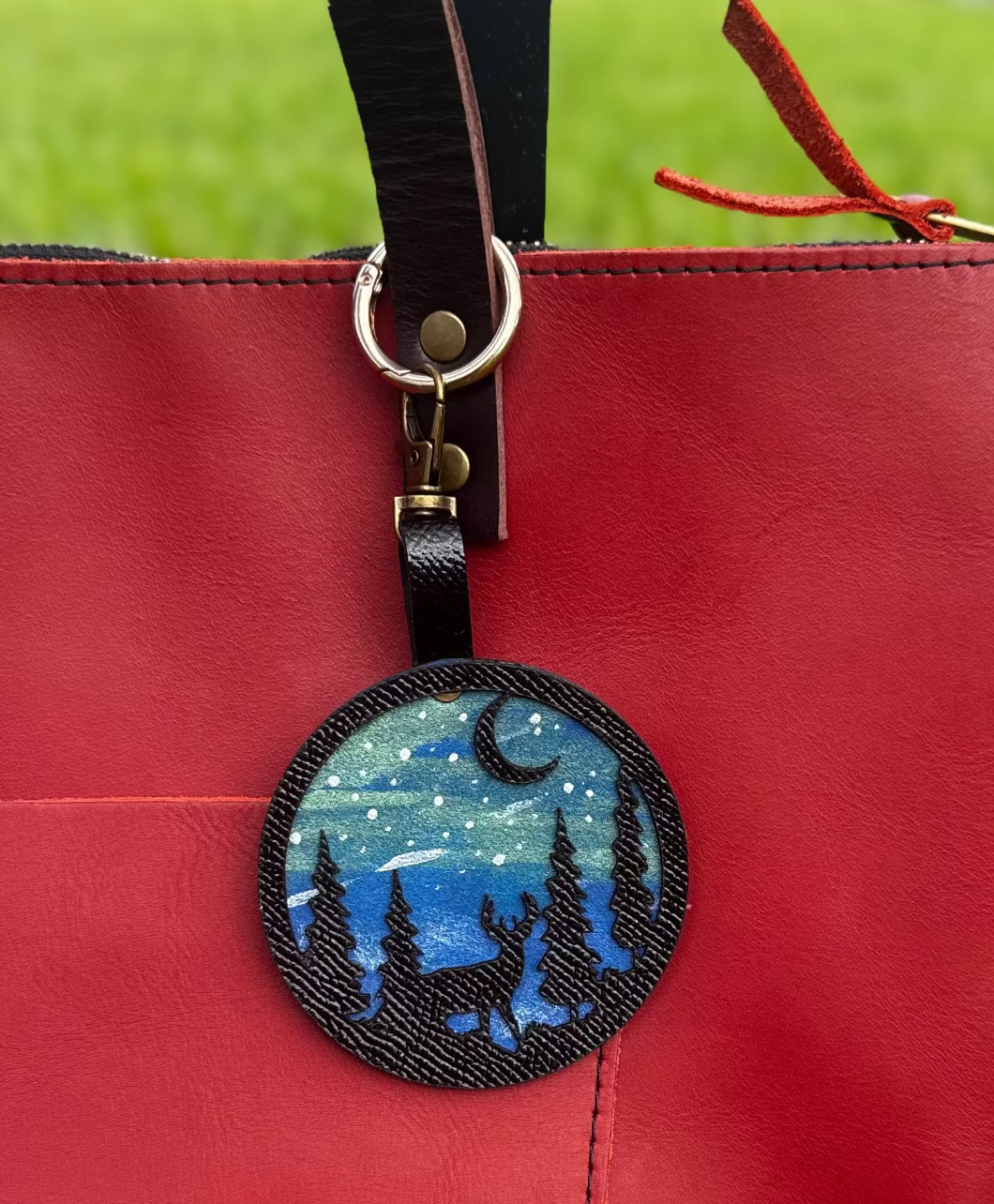 A handpainted leather charm with a starry night landscape, a deer, and a forest design, attached to a red leather bag.