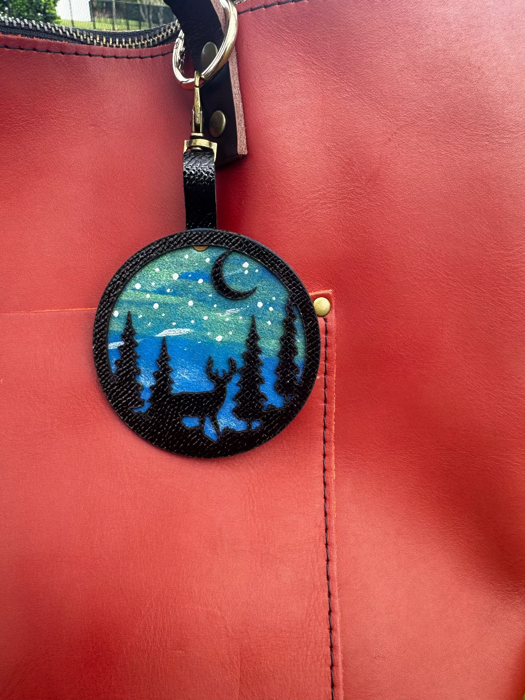 A handpainted leather charm with a starry night landscape, a deer, and a forest design, attached to a red leather bag.