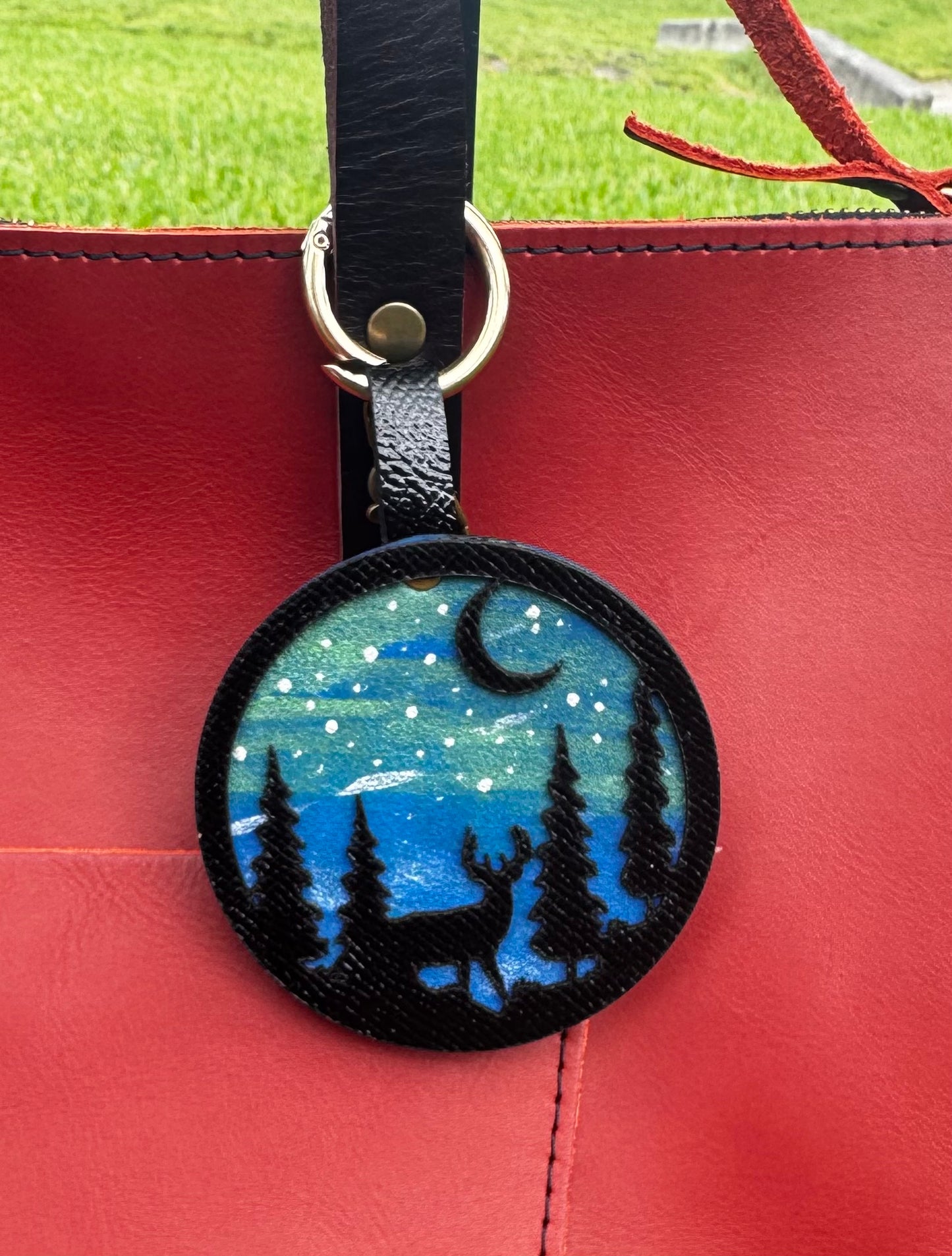 A handpainted leather charm with a starry night landscape, a deer, and a forest design, attached to a red leather bag.