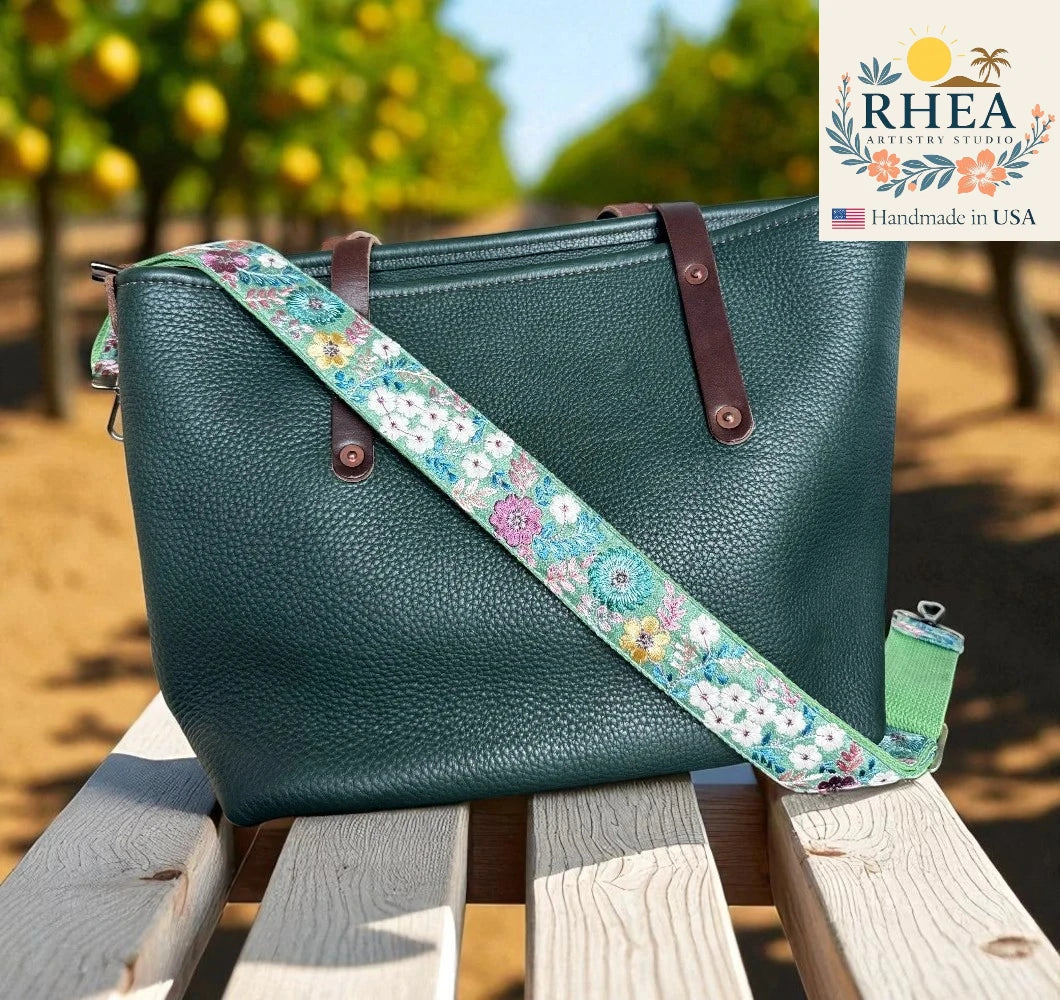 Teal handbag with a floral strap on a wooden surface, with 'RHEA' branding in the corner.