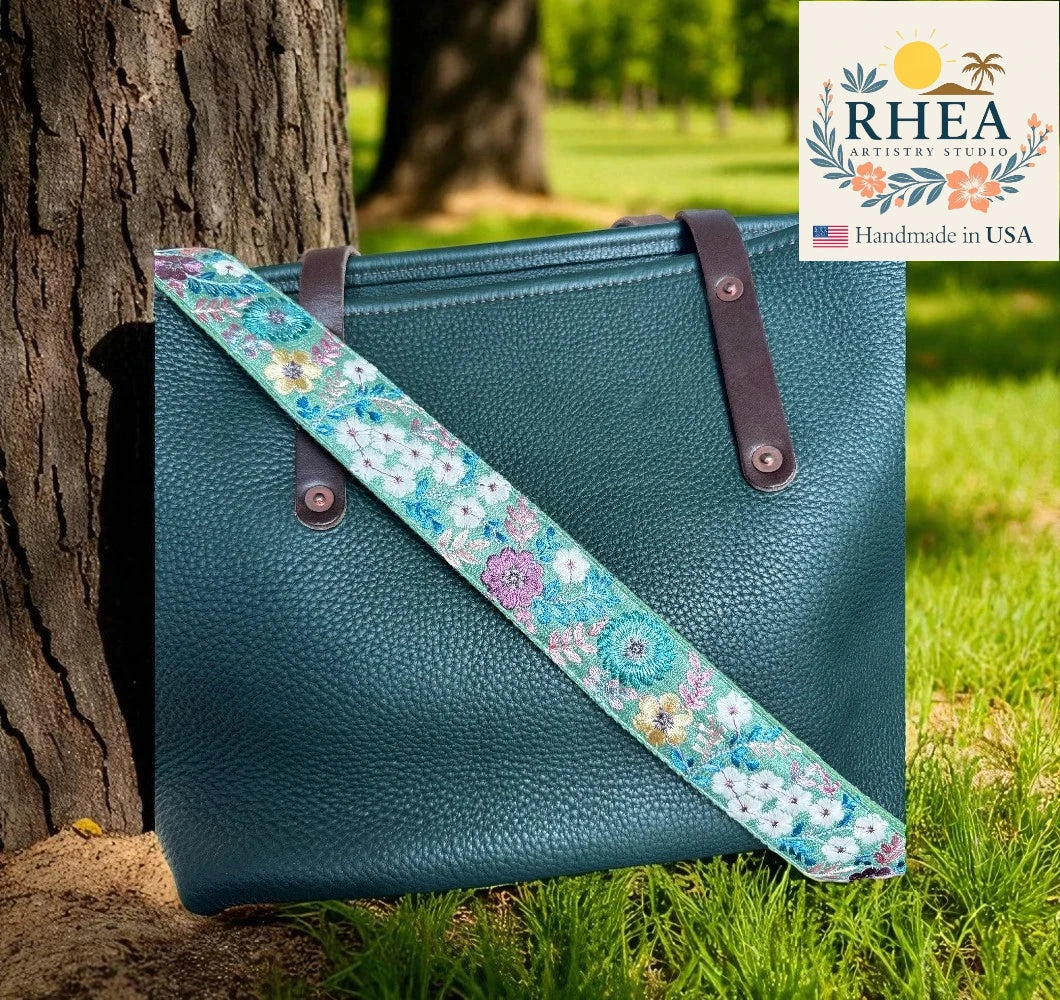 Blue handbag with floral strap leaning against a tree outdoors, RHEA Artistry Studio logo visible.