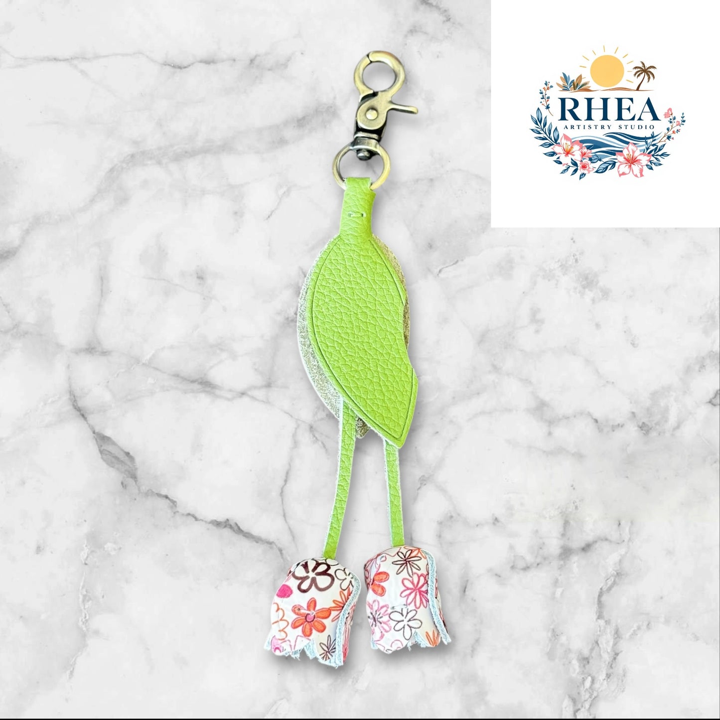 A handmade leather tulip charm with a green leaf and brown floral pattern, attached to a gold lobster clasp on a white marble surface.