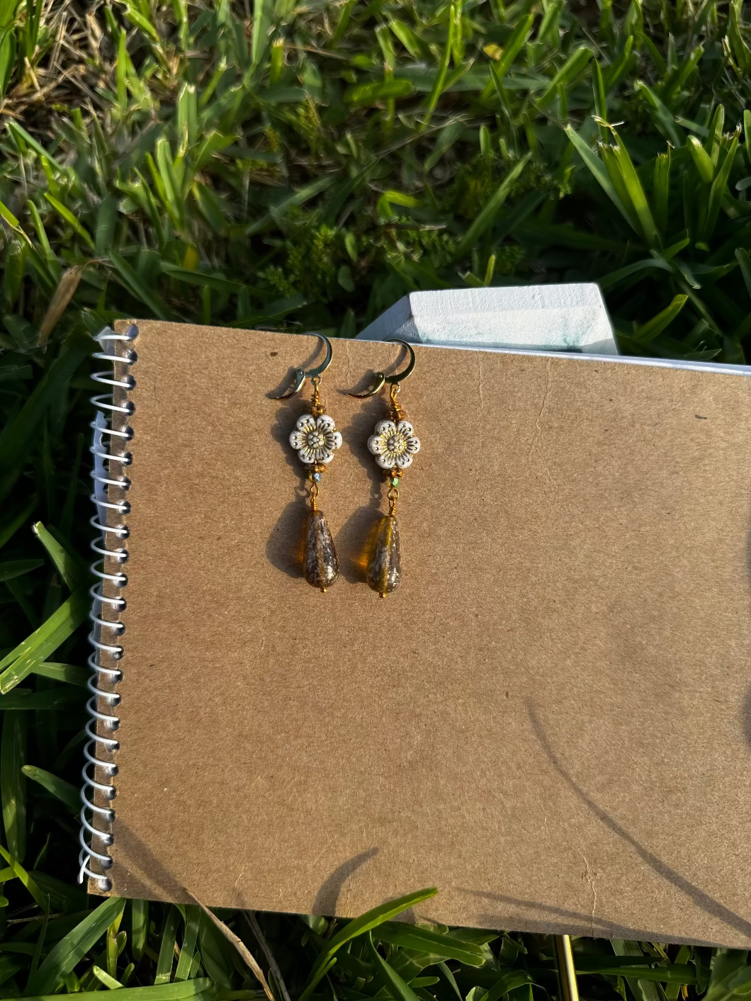 A pair of white hibiscus gold drop earrings with gold plated brass findings, displayed on a notebook.