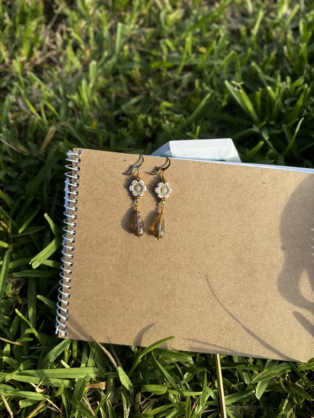 A pair of white hibiscus gold drop earrings with gold plated brass findings, displayed on a notebook.