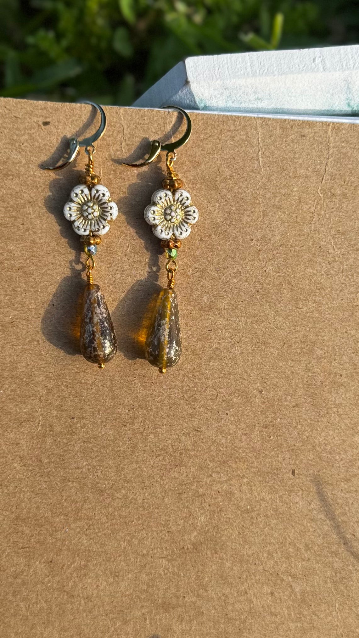A pair of white hibiscus gold drop earrings with gold plated brass findings, displayed on a notebook.