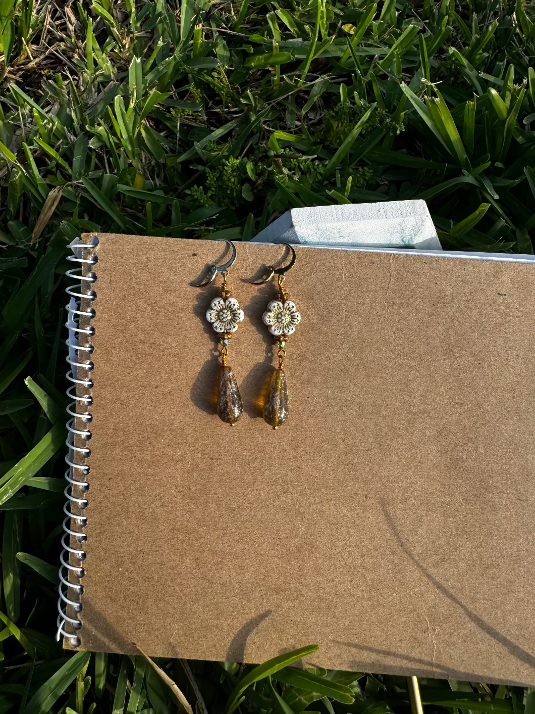 A pair of white hibiscus gold drop earrings with gold plated brass findings, displayed on a notebook.