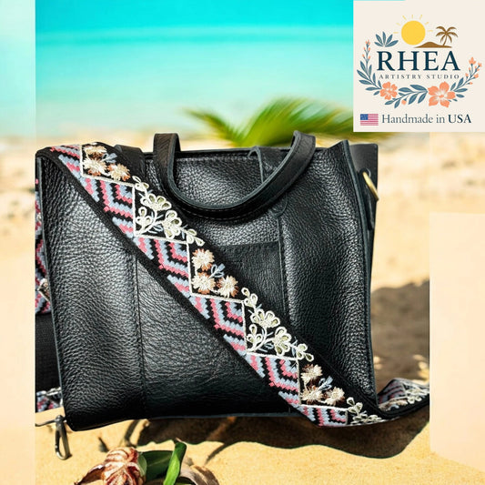 Black handbag with embroidered strap on a beach background, featuring the RHEA Artistry Studio logo.