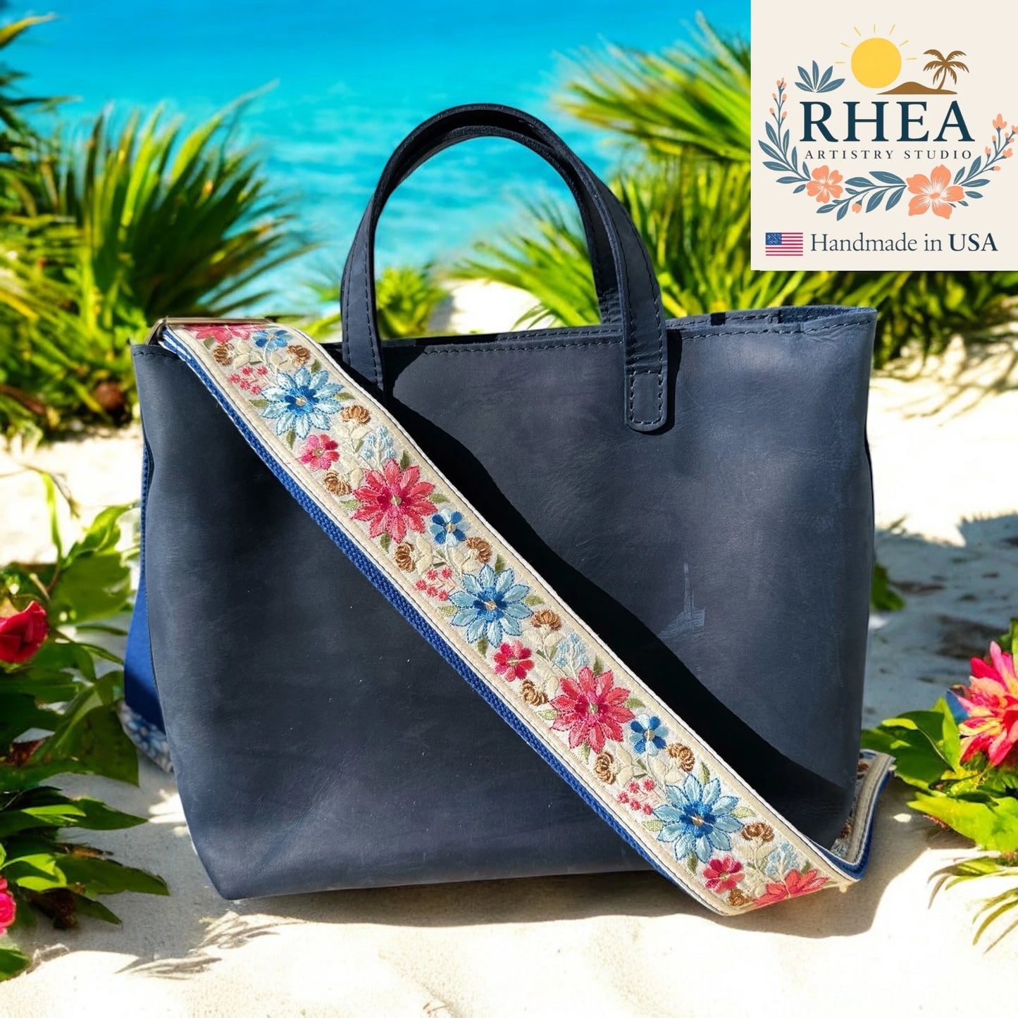 Navy blue handbag with floral purse strap on a beach background, featuring RHEA Artistry Studio logo.