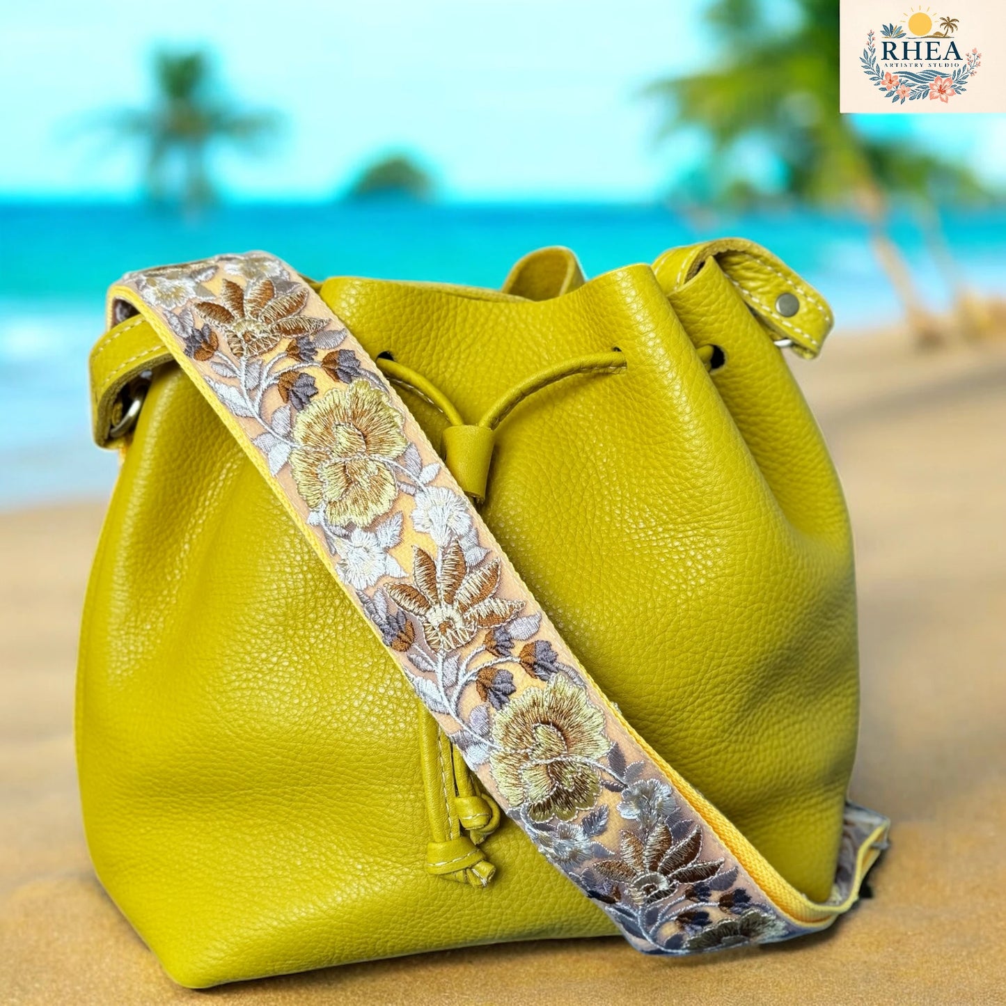 Light yellow handcrafted leather bucket bag with a wide floral embroidered strap, displayed on a tropical beach backdrop — Rose Bloom Collection by Rhea Artistry Studio.