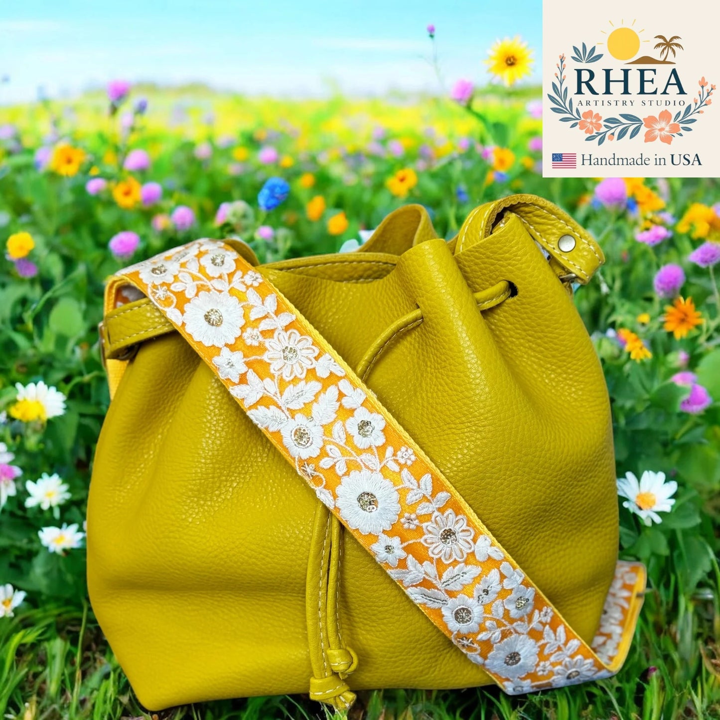 Yellow leather bag with a floral purse strap in a field of flowers, featuring the RHEA Artistry Studio logo.