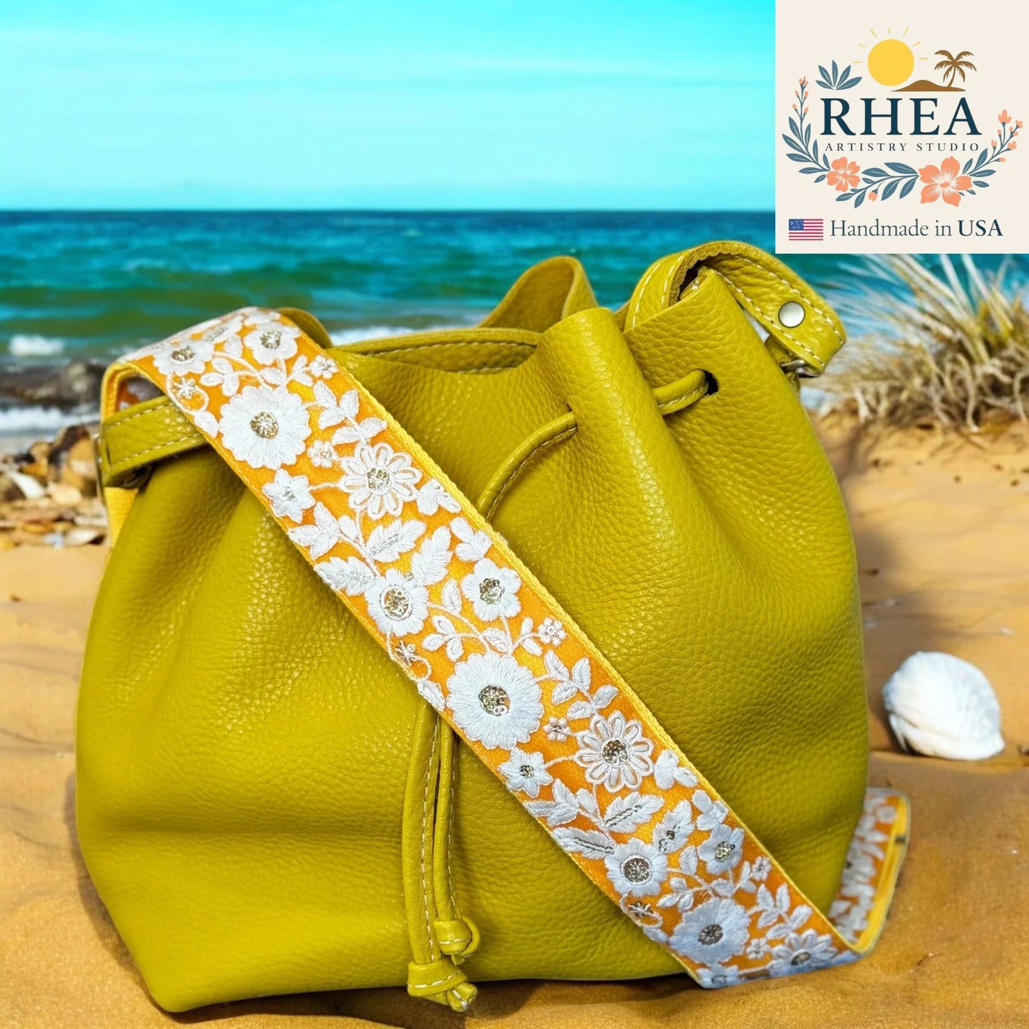 Yellow leather bag with a floral purse strap on a beach, featuring the RHEA Artistry Studio logo.