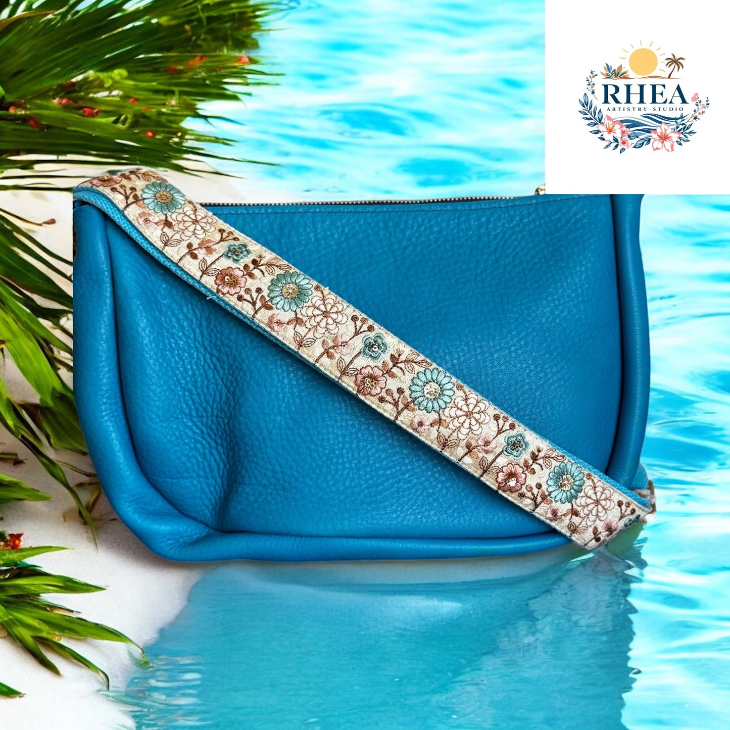 Blue clutch bag with floral strap on a beach setting, featuring the RHEA brand.