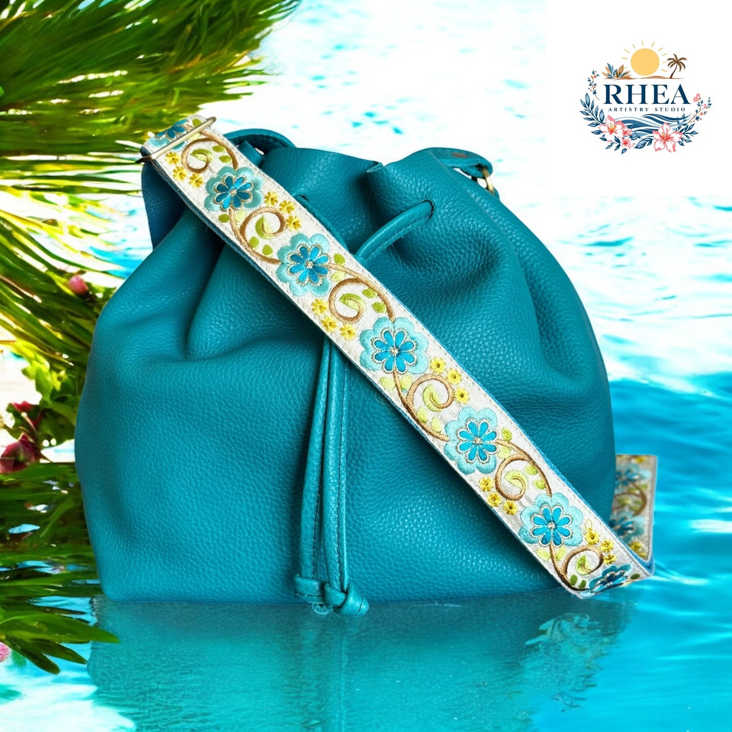 Aqua blue eight-petal floral embroidered strap by Rhea Artistry Studio styled with a teal leather bucket bag. Features intricate high-quality craftsmanship with ethically sourced cotton webbing, perfect as a luxury crossbody strap or camera strap.