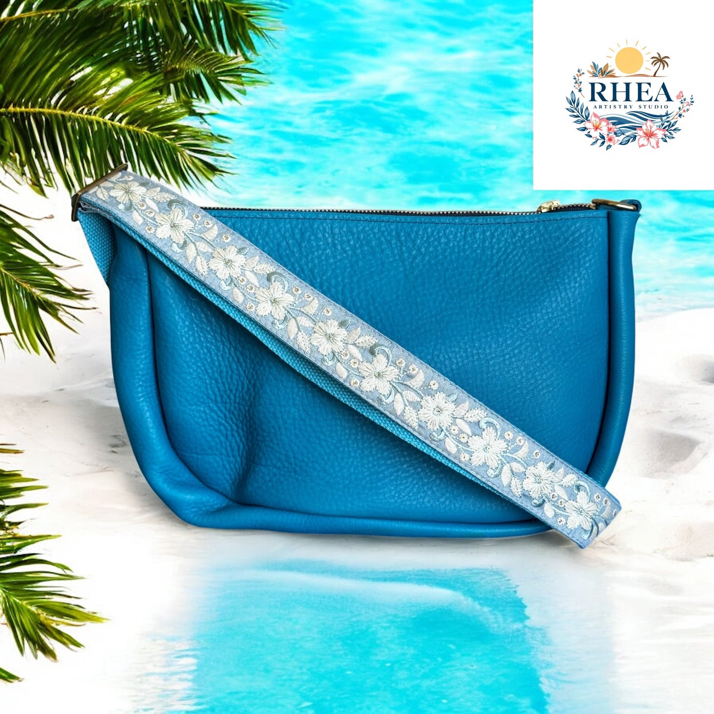 Blue handbag with floral strap on a beach background, featuring the RHEA brand.