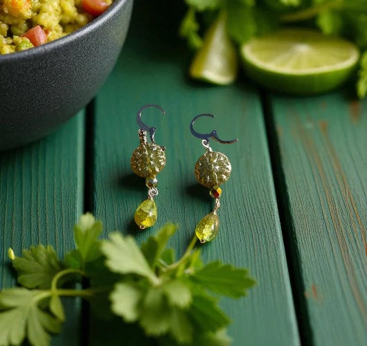 Gold earrings with green stones on a green wooden surface with herbs and a bowl of food.
