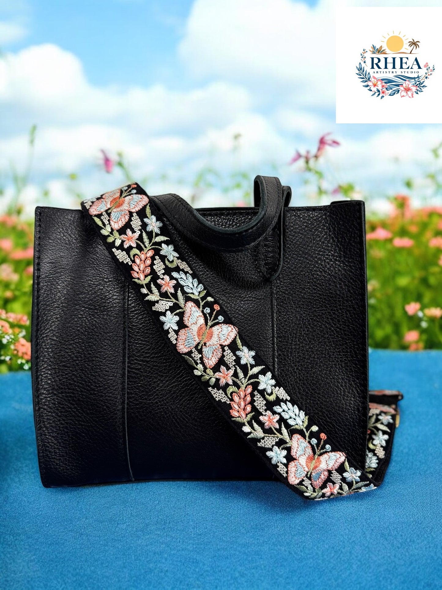 A black purse with a pink and green floral embroidered strap placed on a blue surface with flowers in the background.