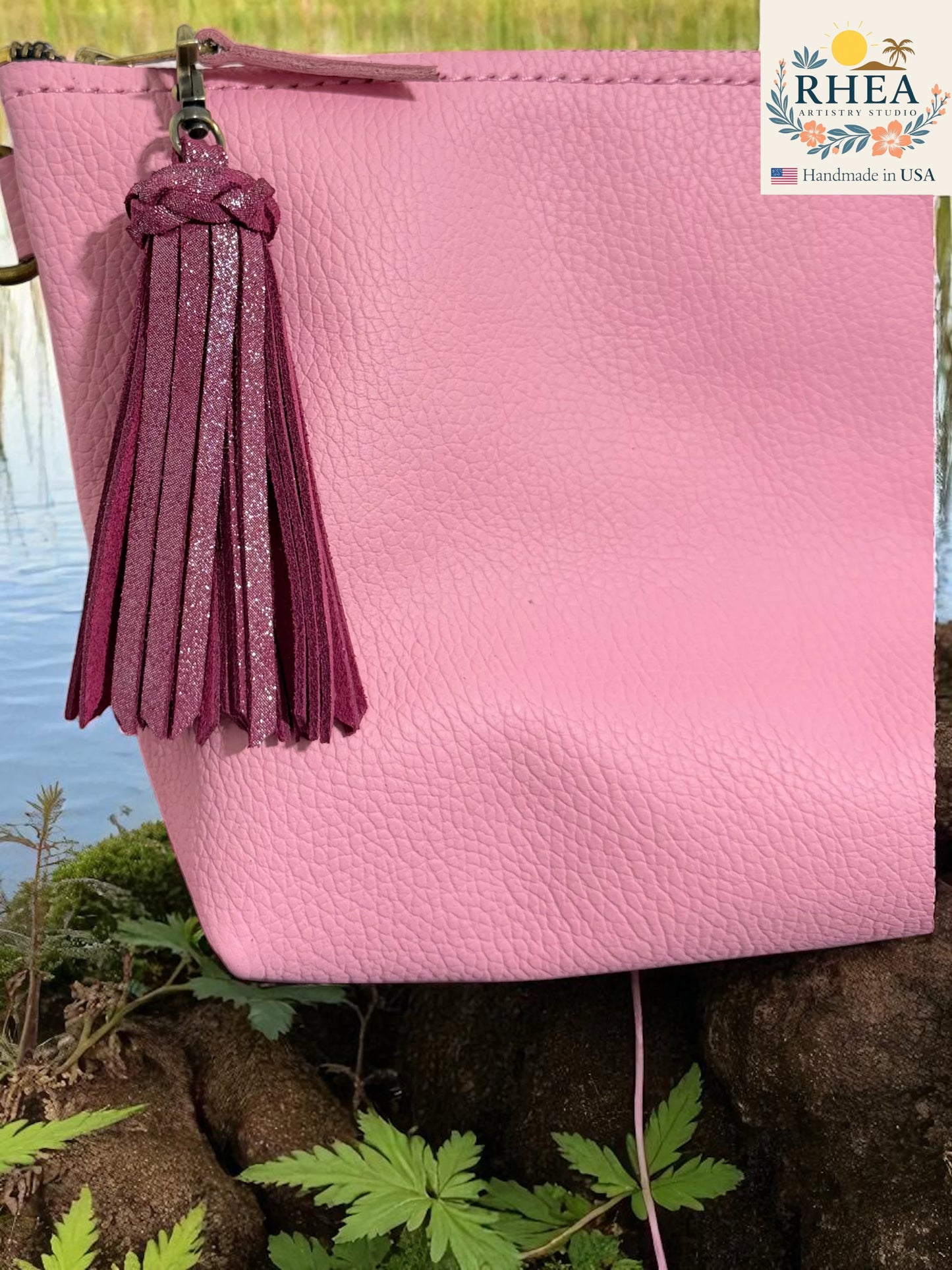 Pink leather clutch with a tassel on a natural background, featuring the RHEEA brand.