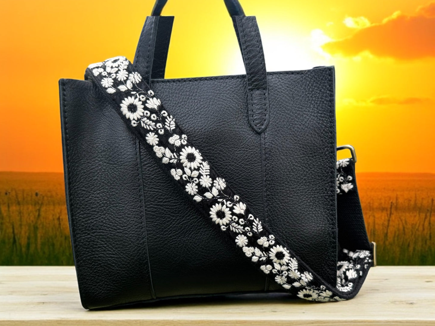 Elegant black fabric crossbody strap with detailed white wildflower embroidery, perfect for handbags and DSLR cameras. Ethically handcrafted with a modern flair against a sunset background
