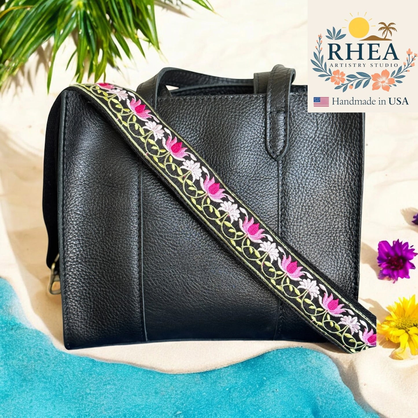 Black handbag with a floral purse strap on a sandy background, featuring the RHEA Artistry Studio logo.