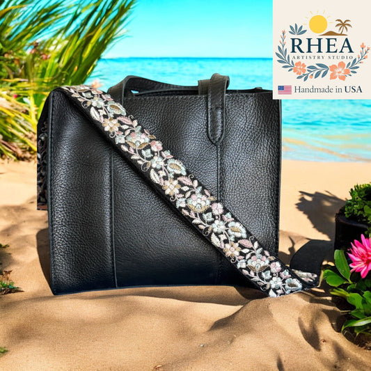 Black handbag with floral purse strap on a beach, featuring RHEA Artistry Studio logo.