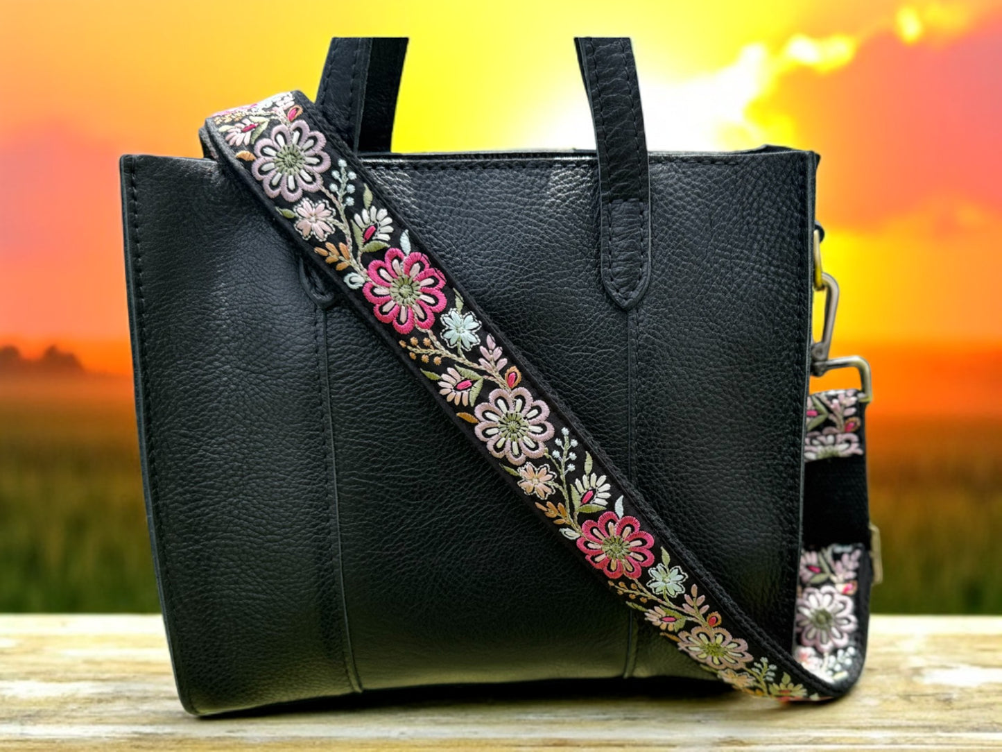 Black bag with vibrant floral crossbody strap featuring pink, ivory, and olive green tones against sunset background.