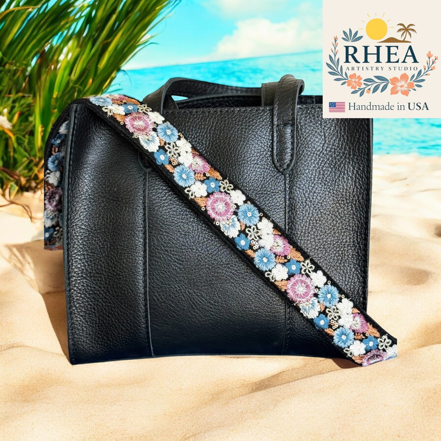 Black handbag with a floral purse strap on a sandy beach, featuring the RHEA Artistry Studio logo.