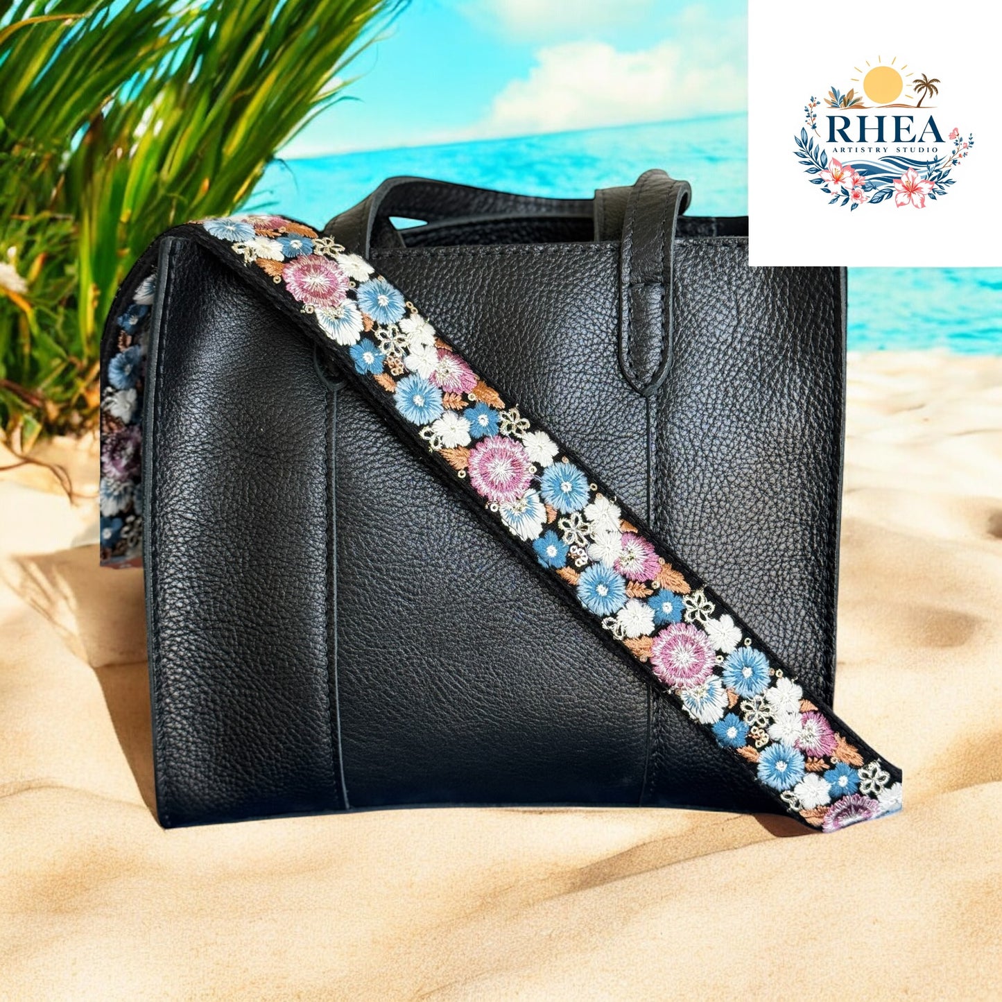 A black leather tote with an embroidered strap featuring blue, pink, and white flowers on a black background, against a tropical sandy beach