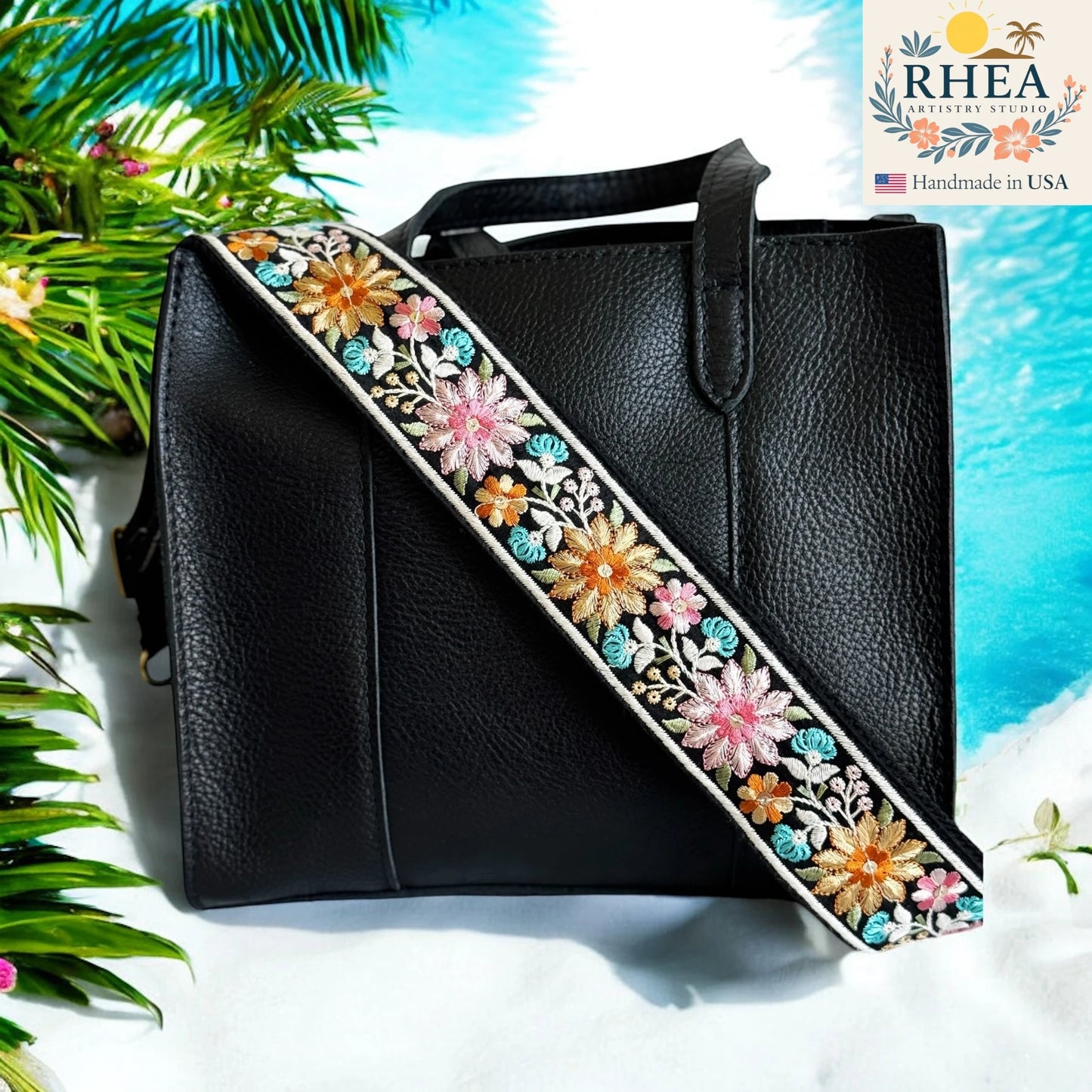 Black handbag with a floral purse strap on a beach-themed background, featuring the RHEA brand.