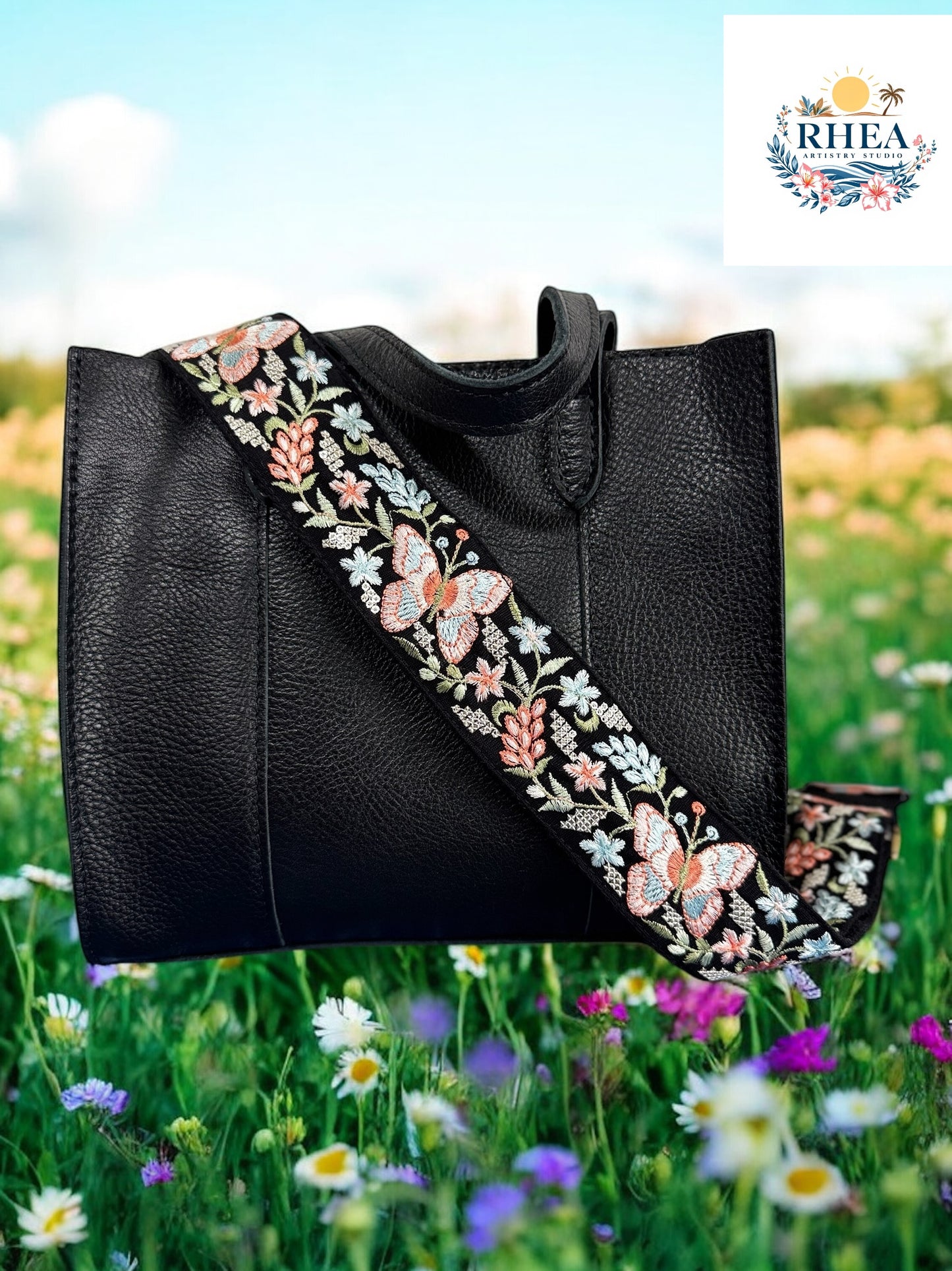 A black purse with a pink and green floral embroidered strap placed on a blue surface with flowers in the background.