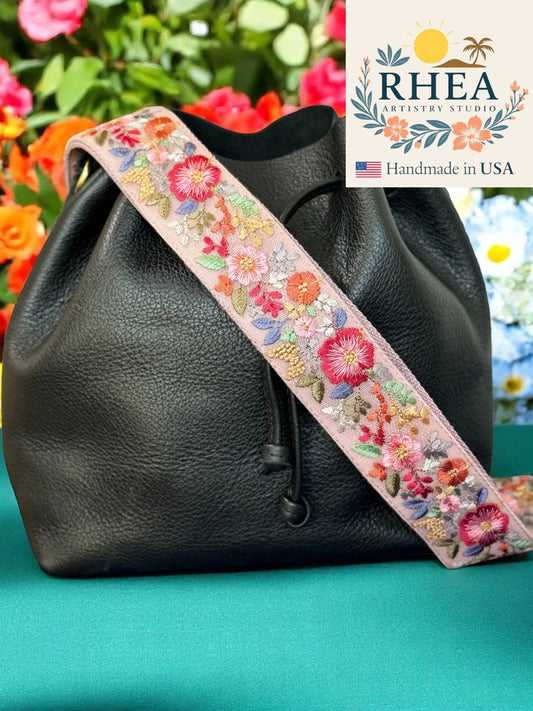 Black leather bag with a floral purse strap on a green background, featuring the RHEA Artistry Studio logo.
